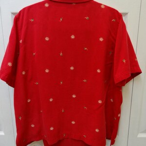 May include: A red short-sleeved button-down shirt with a white embroidered design. The shirt has a collar and a pocket on the left side. The shirt is paired with red trousers.