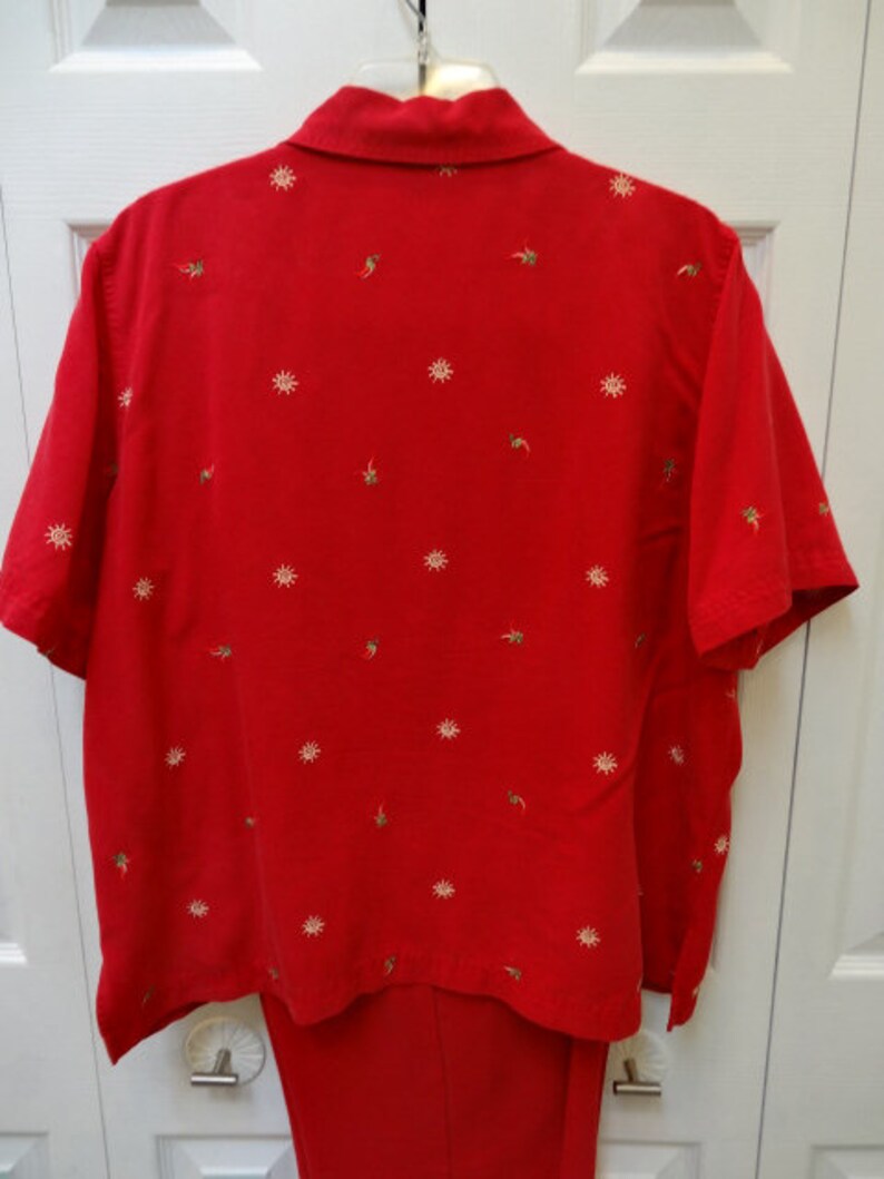 May include: A red short-sleeved button-down shirt with a white embroidered design. The shirt has a collar and a pocket on the left side. The shirt is paired with red trousers.