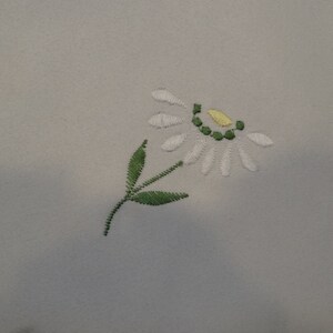 May include: White embroidered flower with a yellow center and green stem and leaves on a white fabric background.