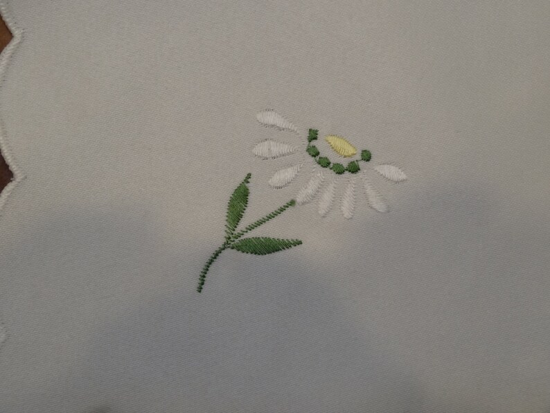 May include: White embroidered flower with a yellow center and green stem and leaves on a white fabric background.