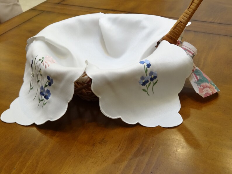 May include: White embroidered linen basket liner with blue and pink flowers. The liner has a scalloped edge and is folded over a wicker basket.