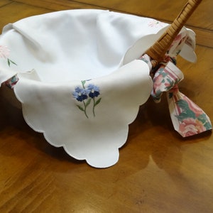 May include: A white linen basket liner with scalloped edges and embroidered flowers. The liner is folded over a wicker basket with a floral patterned fabric handle.