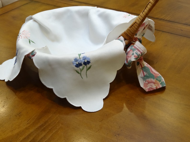 May include: A white linen basket liner with scalloped edges and embroidered flowers. The liner is folded over a wicker basket with a floral patterned fabric handle.