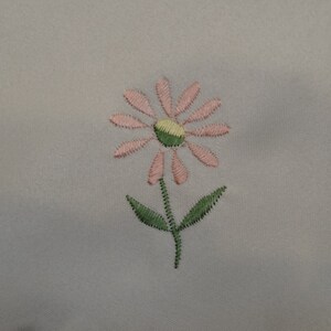 May include: A pink and green embroidered flower design on a white fabric background.