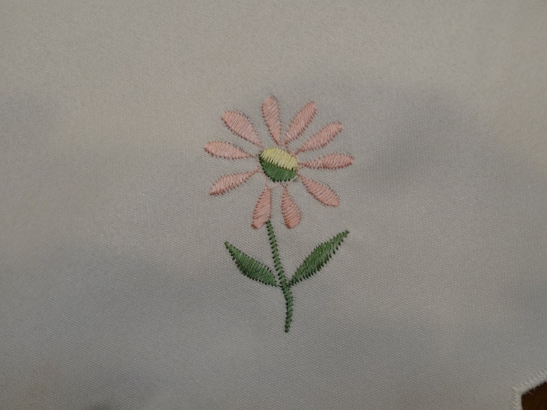 May include: A pink and green embroidered flower design on a white fabric background.