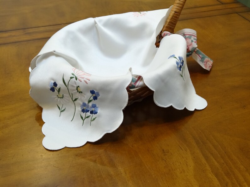 May include: White embroidered fabric basket liner with scalloped edges and floral designs in blue, pink, and green.