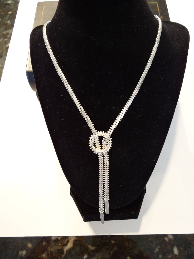 May include: A silver chain necklace with a knot design and a small circle pendant. The necklace is on a black velvet display stand.