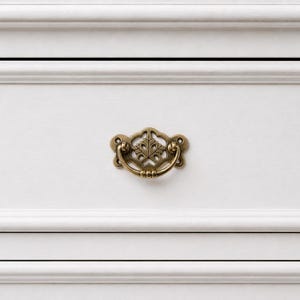 Peut inclure: Gros plan sur un tiroir de commode blanc avec une poignée décorative en laiton. Le tiroir présente un design à panneaux encastrés et fait partie d'un meuble blanc plus grand. La poignée est ornée et ajoute une touche d'élégance.