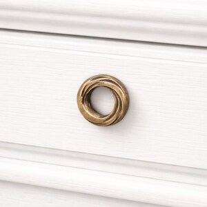 Peut inclure: Gros plan d'un bouton de meuble circulaire de couleur bronze avec un design torsadé texturé. Le bouton est monté sur une surface en bois blanc avec des panneaux horizontaux. Le design du bouton ajoute une touche d'élégance.