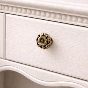 Peut inclure: Gros plan sur un tiroir en bois blanc avec un bouton de meuble en laiton antique. Le bouton est rond avec un motif floral orné. Le tiroir fait partie d'un meuble plus grand.