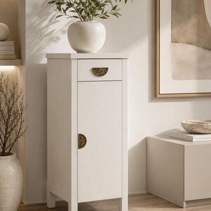 Peut inclure: Un meuble en bois blanc avec un seul tiroir et une porte, orné de poignées en laiton. Un vase rond de couleur claire avec des branches vertes feuillues est posé sur le dessus. Le meuble est de conception étroite et verticale.