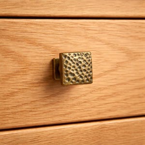Peut inclure: Gros plan sur un bouton de meuble carré en laiton martelé. Le bouton est monté sur un tiroir en bois clair. Le bouton présente une surface texturée avec une série de petites indentations irrégulières.