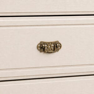 Peut inclure: Gros plan sur un tiroir de commode crème avec une poignée décorative en laiton. Le tiroir présente un design de panneau encastré et une surface légèrement texturée. La poignée est ornée d'un design détaillé d'inspiration vintage.