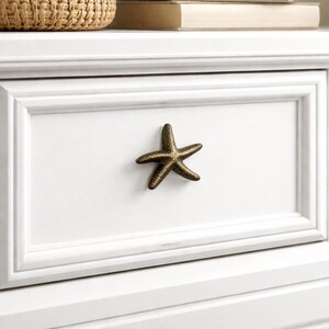 Peut inclure: Poignée de tiroir en forme d'étoile de mer de couleur bronze, fixée sur une armoire blanche. L'étoile de mer texturée à cinq branches ajoute un thème côtier. L'armoire présente un design à panneaux encastrés.