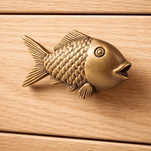 Peut inclure: Une poignée de meuble en forme de poisson de couleur bronze. La quincaillerie décorative présente un motif détaillé d'écailles, d'ailerons et d'une bouche ouverte. La poignée est montée sur une façade de tiroir en bois clair.