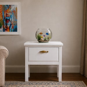 Peut inclure: Une table de chevet blanche avec un tiroir et une poignée dorée. Un bocal à poissons rond en verre avec des poissons colorés est posé dessus. Une peinture abstraite est accrochée au mur, et un canapé beige est en arrière-plan.