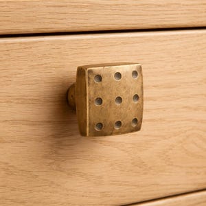 Peut inclure: Un bouton de tiroir carré en laiton antique avec neuf indentations circulaires disposées comme un dé. Le bouton est monté sur un tiroir en bois de couleur claire, mettant en valeur le design et la finition de la quincaillerie. C'est un élément décoratif.