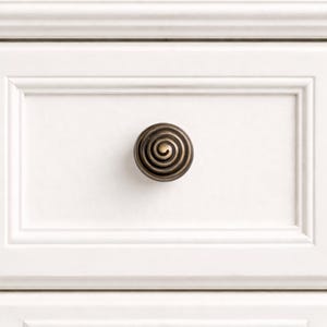Peut inclure: Gros plan sur un tiroir de meuble blanc avec un bouton décoratif en spirale de couleur bronze. Le bouton est centré sur le tiroir, qui présente un motif de panneau encastré. Le meuble semble être peint en blanc.