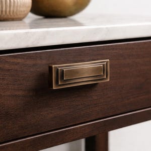 Peut inclure: Gros plan sur un tiroir en bois brun foncé avec une poignée rectangulaire en laiton antique. Le tiroir fait partie d'un meuble plus grand, avec un comptoir en marbre blanc visible au-dessus.