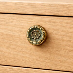 Peut inclure: Gros plan sur un bouton de tiroir décoratif en laiton avec un motif floral. Le bouton rond présente un motif floral en relief détaillé et une patine vert foncé. Il est monté sur une surface en bois clair.