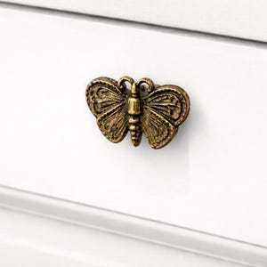 Peut inclure: Un bouton de meuble en forme de papillon de couleur bronze. Le bouton est monté sur une surface blanche. Le papillon présente des motifs d'ailes détaillés et un corps texturé, ajoutant une touche décorative aux meubles.