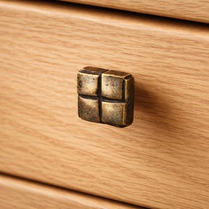 Peut inclure: Gros plan d'un bouton de tiroir carré en laiton antique avec un motif en forme de croix. La quincaillerie est montée sur une façade de tiroir en bois clair, mettant en valeur le grain naturel du bois.