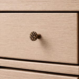 Peut inclure: Gros plan sur un tiroir de meuble en bois clair avec un bouton décoratif rond de couleur bronze. Le tiroir fait partie d'un meuble plus grand avec le grain du bois visible. Le bouton a un motif quadrillé.