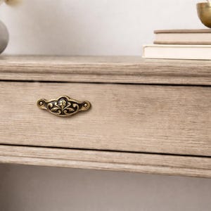 Peut inclure: Un tiroir en bois brun clair doté d'une poignée décorative en laiton antique avec un motif floral orné. Le tiroir fait partie d'un meuble en bois avec une texture de grain de bois visible et une forme rectangulaire horizontale.