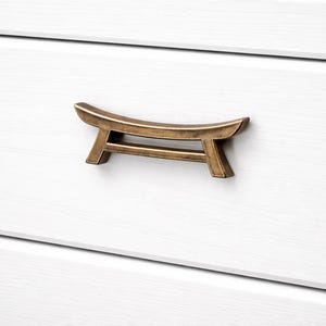 Peut inclure: Une poignée de tiroir de couleur bronze au design incurvé unique. La quincaillerie est montée sur un tiroir blanc, offrant un contraste de couleur et de texture. La poignée a une esthétique minimaliste d'inspiration asiatique.