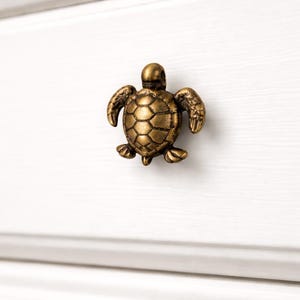 Peut inclure: Bouton de meuble en forme de tortue de couleur bronze. Le bouton est fixé sur une porte d'armoire blanche. La tortue présente des détails, notamment une carapace texturée et des membres en forme de nageoires. Décoratif.