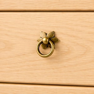 Peut inclure: Gros plan sur une poignée de tiroir en forme de fleur en laiton avec une poignée en anneau. La poignée est montée sur une surface en bois clair, mettant en valeur le grain du bois. La quincaillerie ajoute un charme vintage aux meubles.