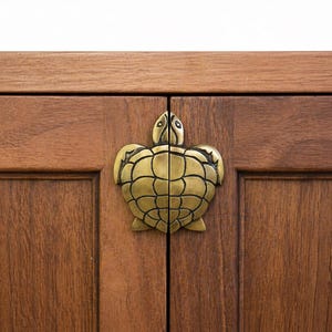 Peut inclure: Gros plan sur une armoire en bois avec une poignée en forme de tortue en laiton. Les portes de l'armoire sont de couleur marron chaud, et la poignée en forme de tortue est centrée, ajoutant une touche décorative.