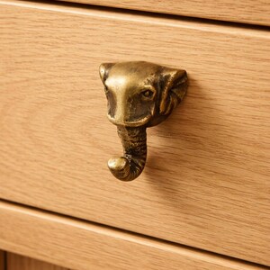 Peut inclure: Poignée de tiroir en forme de tête d'éléphant en laiton, fixée sur une armoire en bois clair. La poignée est détaillée avec les traits d'un éléphant, dont les défenses et une trompe enroulée. Le grain du bois est visible.