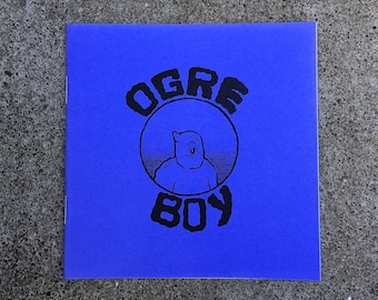 Può includere: Un libretto blu con il testo "OGRE BOY" in lettere nere. Un'illustrazione in bianco e nero di un orco con un occhio solo è all'interno di un cerchio.