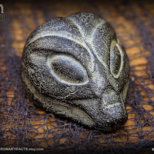 May include: A detailed, dark gray sculpture of an alien head with large, oval eyes and textured skin. The sculpture is set against a dark, textured background. The Etsy shop name, Landron Artifacts, is visible in the image.