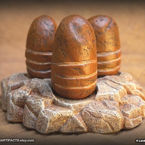 May include: Three brown, bullet-shaped sculptures with horizontal grooves, resting on a textured, rock-like base. The sculptures have a weathered appearance, suggesting an aged or antique aesthetic. The base is a light brown color.