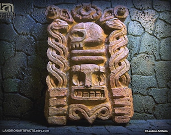 May include: A rectangular stone-colored wall plaque featuring a central human-like face mask topped with a skull. Two intertwined snakes flank the sides, with smaller skulls at the top corners. The text Landron Artifacts appears in the corner.