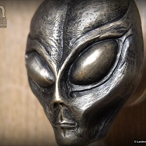 May include: A silver-colored alien head sculpture with a detailed face and large eyes. The sculpture is mounted on a wooden surface.