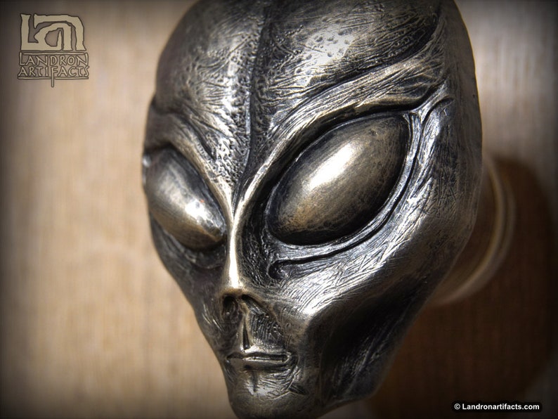 May include: A silver-colored alien head sculpture with a detailed face and large eyes. The sculpture is mounted on a wooden surface.