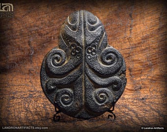 May include: A dark gray, oval-shaped artifact with intricate, swirling patterns and a textured surface. The design features symmetrical, stylized floral and spiral motifs. The artifact is displayed on a small, black metal stand. The Etsy shop URL is visible at the bottom.