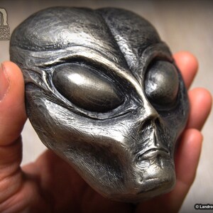 May include: A silver-colored alien head sculpture with large, almond-shaped eyes. The sculpture is detailed with textured skin and a slightly open mouth.
