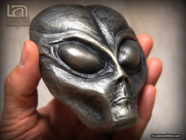 May include: A silver-colored alien head sculpture with large, almond-shaped eyes. The sculpture is detailed with textured skin and a slightly open mouth.