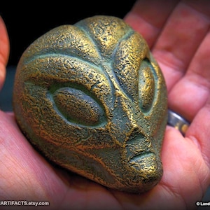 May include: A bronze-colored alien head sculpture with a textured surface. The sculpture has large oval eyes and a small mouth. The alien head is held in a person's hand. The sculpture has a weathered, antique appearance.