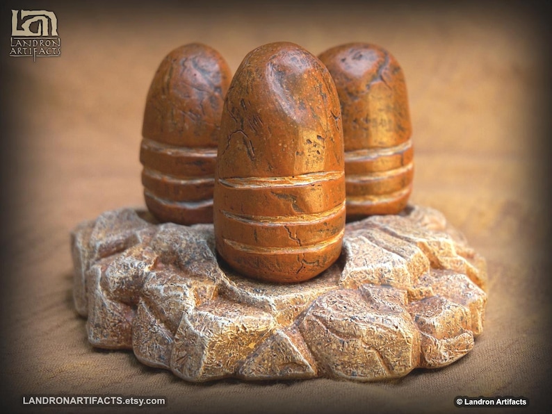 May include: Three brown, bullet-shaped sculptures with horizontal grooves, resting on a textured, rock-like base. The sculptures have a weathered appearance, suggesting an aged or antique aesthetic. The base is a light brown color.