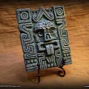 May include: A rectangular, weathered-looking stone carving of a stylized face with a protruding tongue, set on a black metal stand. The carving features intricate geometric patterns and is in shades of gray and green.
