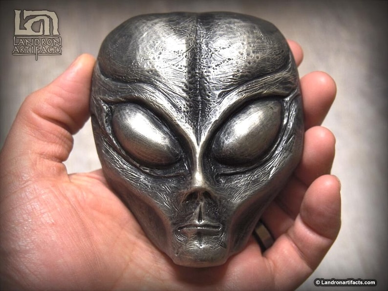 May include: A silver-colored alien head sculpture with large, almond-shaped eyes and a detailed, textured surface.