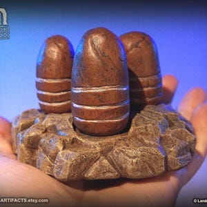 May include: Three brown, egg-shaped sculptures with white bands, set on a textured, rock-like base. The sculptures are held in a hand, with a blue background. The Etsy shop name, Landron Artifacts, is visible.