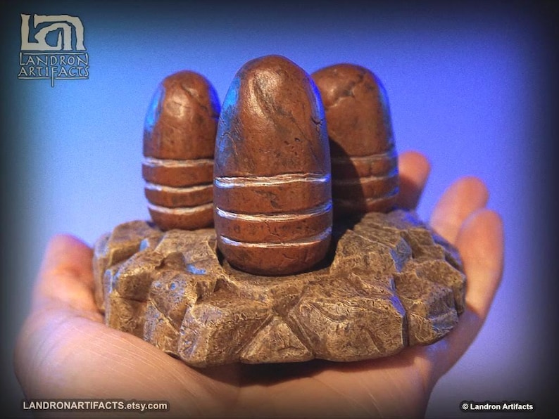 May include: Three brown, egg-shaped sculptures with white bands, set on a textured, rock-like base. The sculptures are held in a hand, with a blue background. The Etsy shop name, Landron Artifacts, is visible.