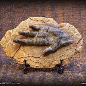 May include: A sculpted hand, detailed with wrinkles and texture, is set on a tan-colored, rock-like base. The hand is a dark gray color. The base is mounted on a black metal stand. The Etsy shop name, Landron Artifacts, is visible.
