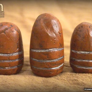 May include: Three brown, egg-shaped artifacts with horizontal silver bands. The objects have a textured surface and a weathered appearance. The artifacts are displayed on a tan surface. The Landron Artifacts logo is in the upper left corner.
